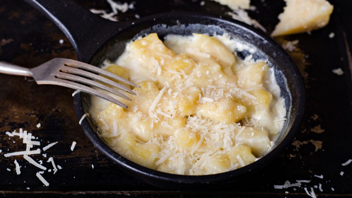 Pariser Gnocchi in cremiger Sahnesauce mit geriebenem Parmesan in einer schwarzen Pfanne und daneben eine Gabel auf dunklem Hintergrund.