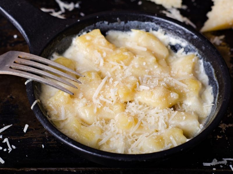 Pariser Gnocchi in cremiger Sahnesauce mit geriebenem Parmesan in einer schwarzen Pfanne und daneben eine Gabel auf dunklem Hintergrund.