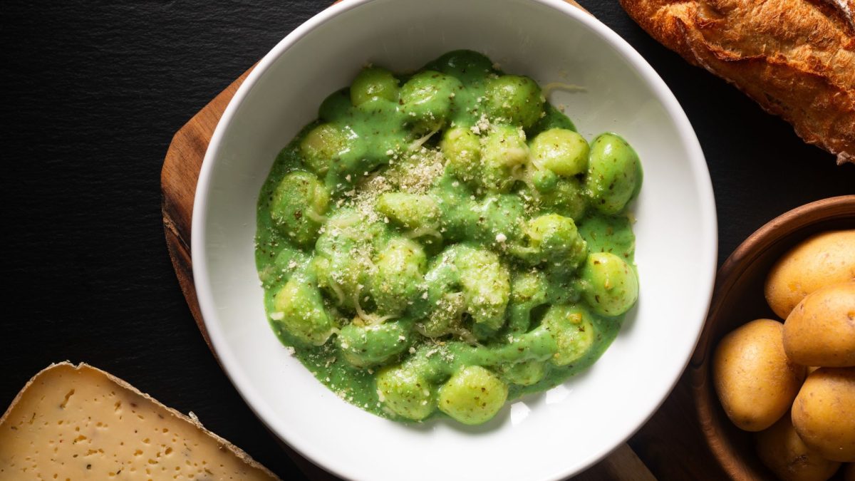 Gnocchi in einer grünen Pesto-Sauce, bestreut mit Parmesan, serviert in einer weißen Schüssel auf schwarzem Untergrund mit Baguette und Kartoffeln.