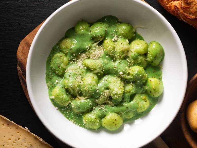 Gnocchi in einer grünen Pesto-Sauce, bestreut mit Parmesan, serviert in einer weißen Schüssel auf schwarzem Untergrund mit Baguette und Kartoffeln.