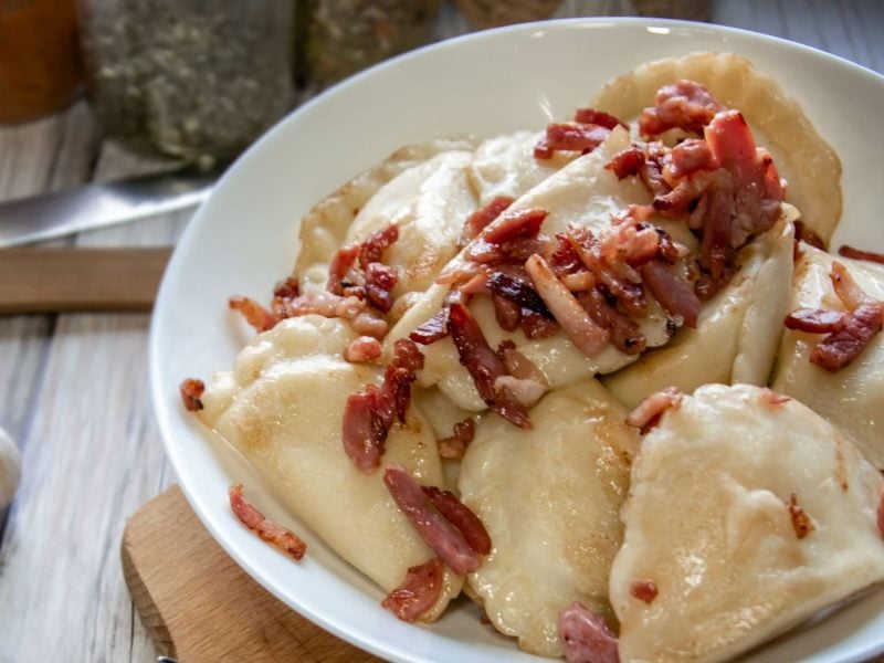 Weiße Schüssel mit polnischen Pierogi, garniert mit gebratenen Speckstücken auf hell gemustertem Tisch.