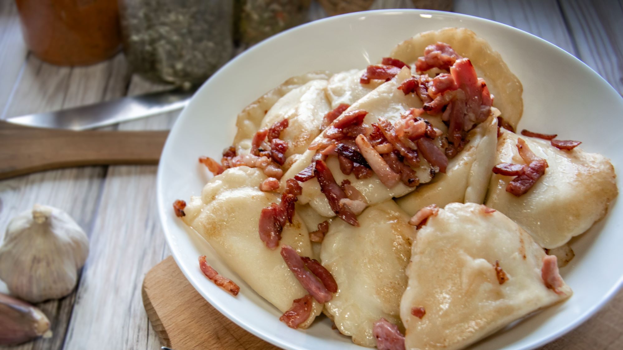 Weiße Schüssel mit polnischen Pierogi, garniert mit gebratenen Speckstücken auf hell gemustertem Tisch.