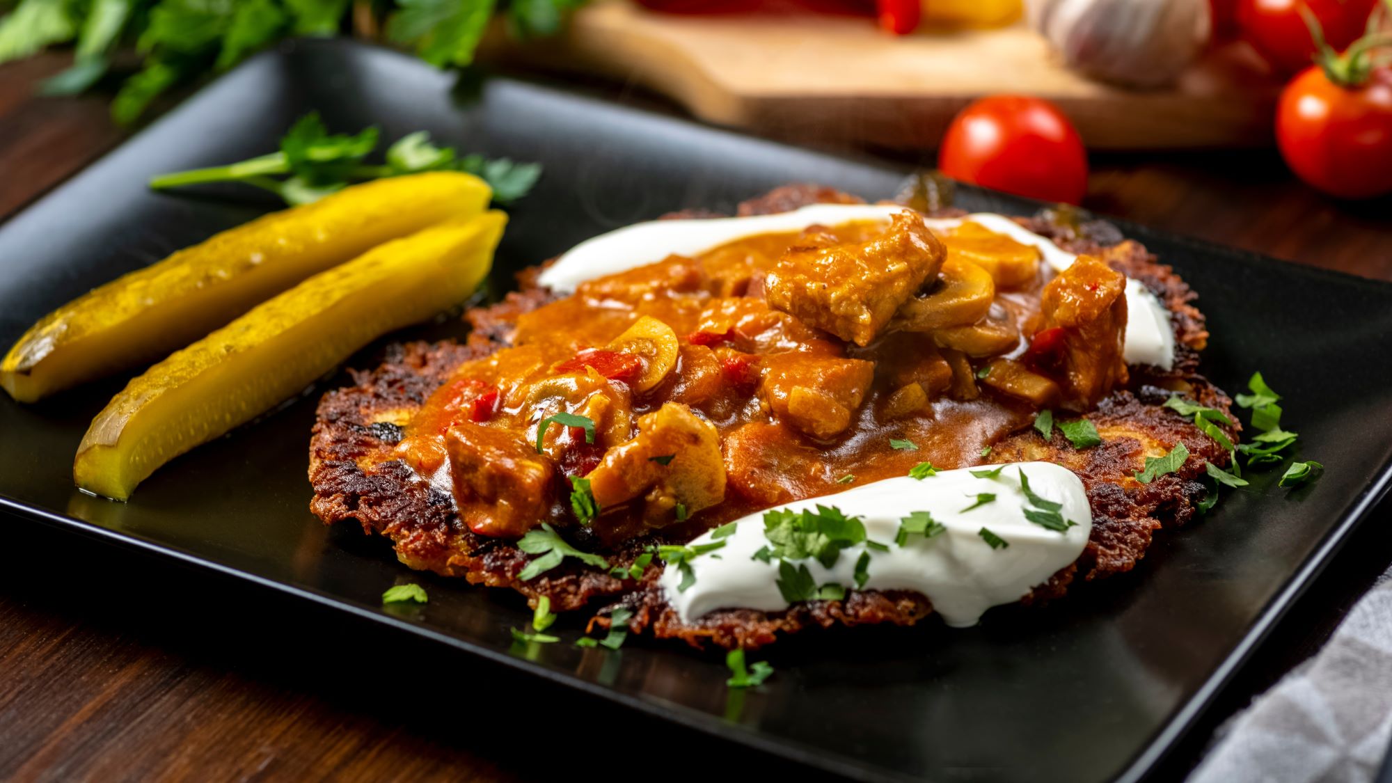 Polnische Kartoffelpuffer "Placek" mit Gulasch und saurer Sahne, dazu zwei Gewürzgurken auf schwarzem Teller, umgeben von frischen Kräutern.