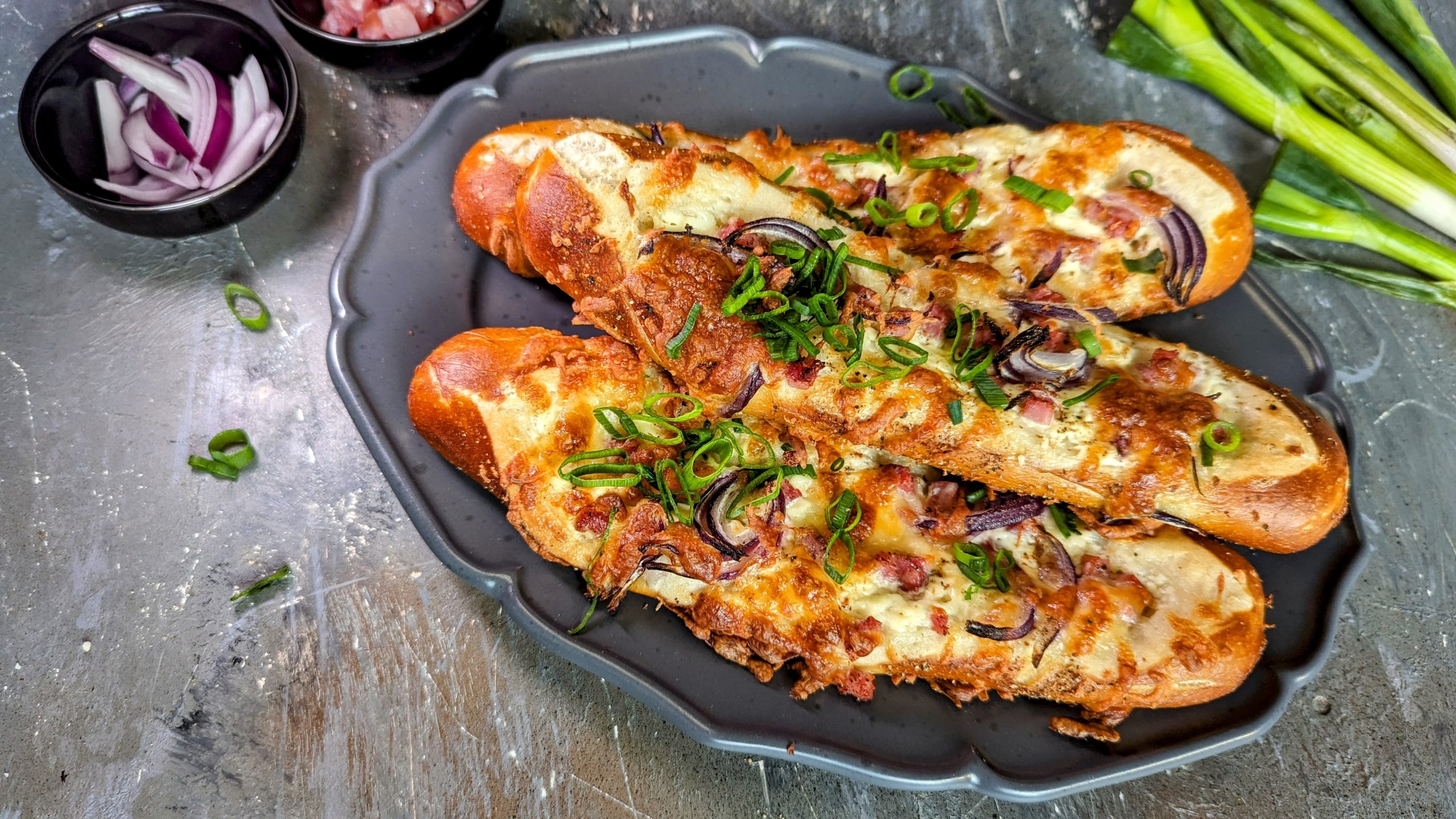 Mehrere Laugenstangen-Flammkuchen auf einem grauen Teller un dgrauem Steinuntergrund direkt von oben fotografiert. Über dme Teller noch diverse Zutaten, die zur Zubereitung nötig sind.