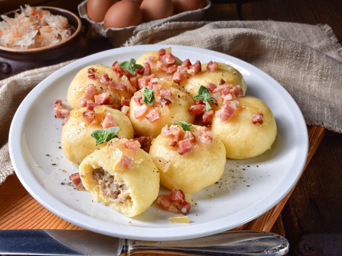 Aufgeschnittene Kartoffelklöße aus Polen namens "Pyzy" mit Fleischfüllung, belegt mit knusprigen Speckwürfeln und Kräutern auf weißem Teller, daneben Sauerkraut.