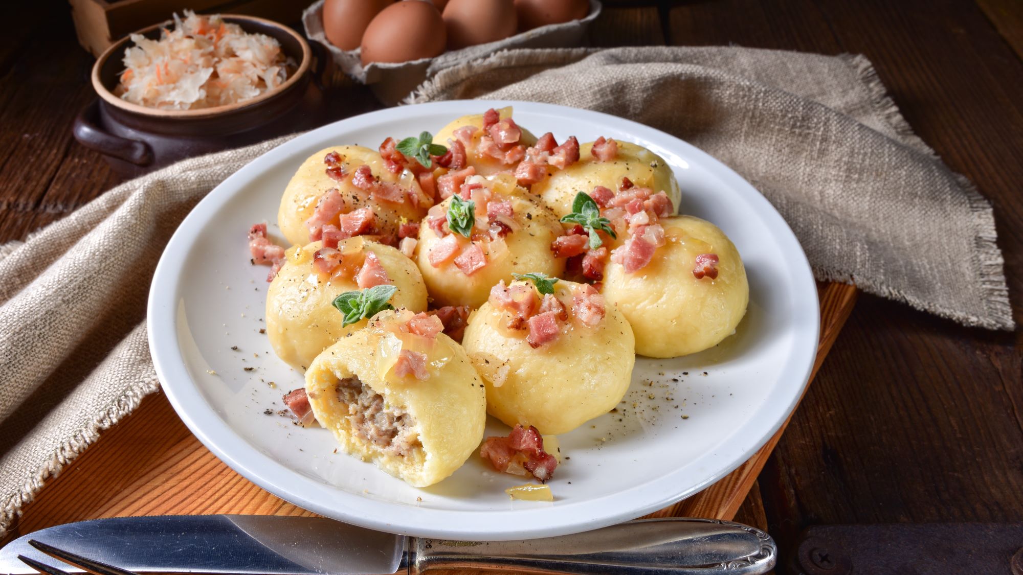 Aufgeschnittene Kartoffelklöße aus Polen namens "Pyzy" mit Fleischfüllung, belegt mit knusprigen Speckwürfeln und Kräutern auf weißem Teller, daneben Sauerkraut.