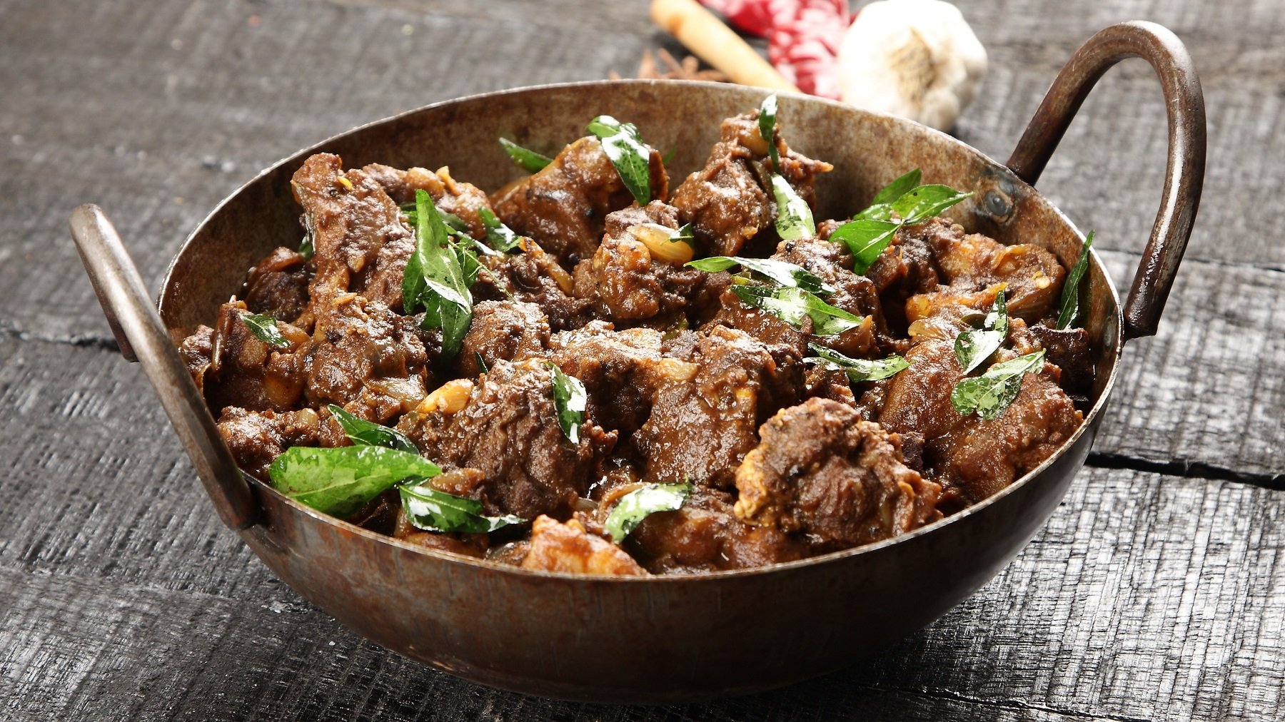 Ein kupferner Topf mit dem Rendang auf einem rustikalen Holztisch. Im Hintergrund Knoblauch, Chili und Zitronengras.