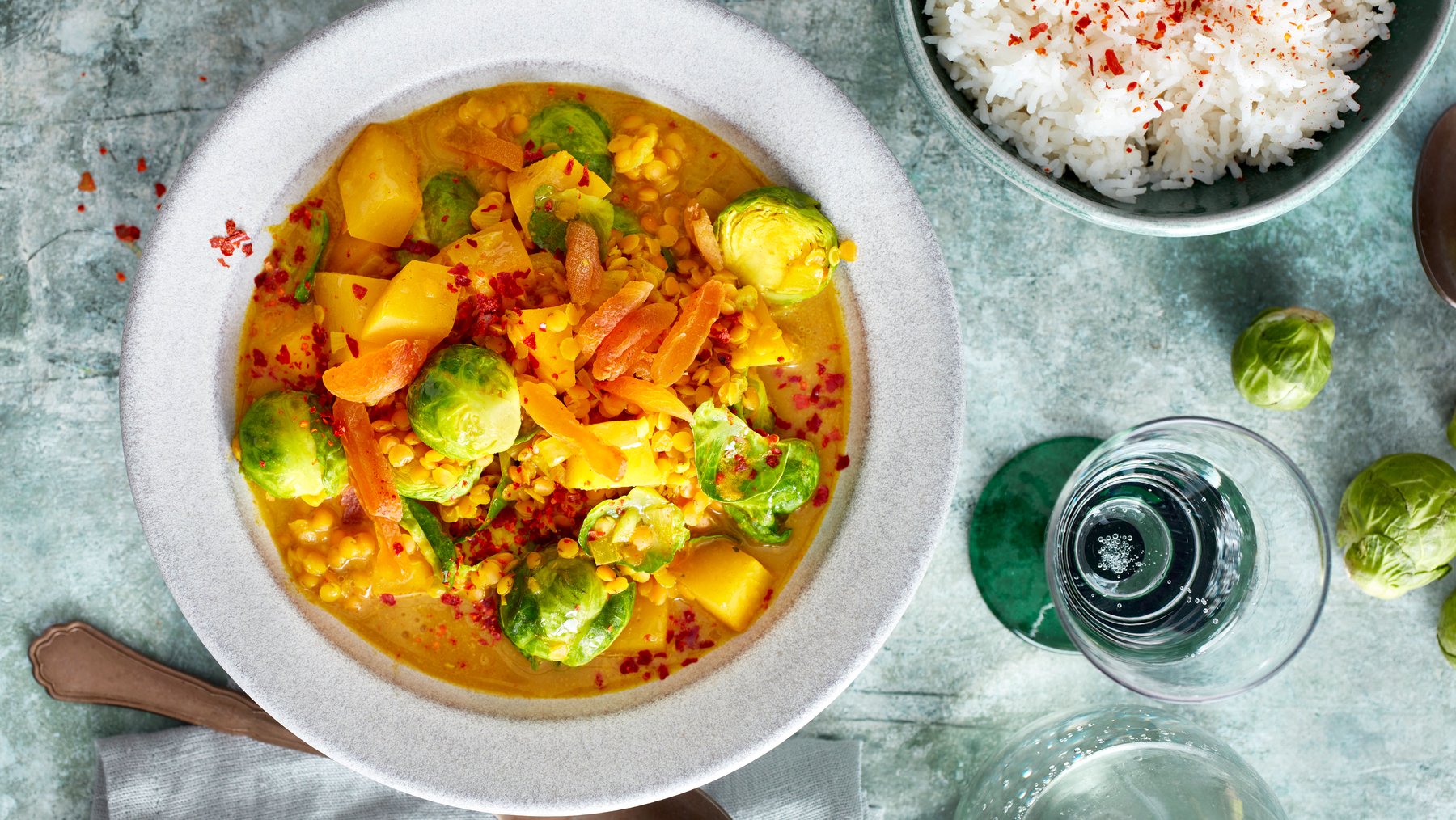 Draufsicht: Rosenkohl-Steckrüben-Curry in einem tiefen Teller, daneben Besteck, Wasserglas und Reis in einer Schüssel.