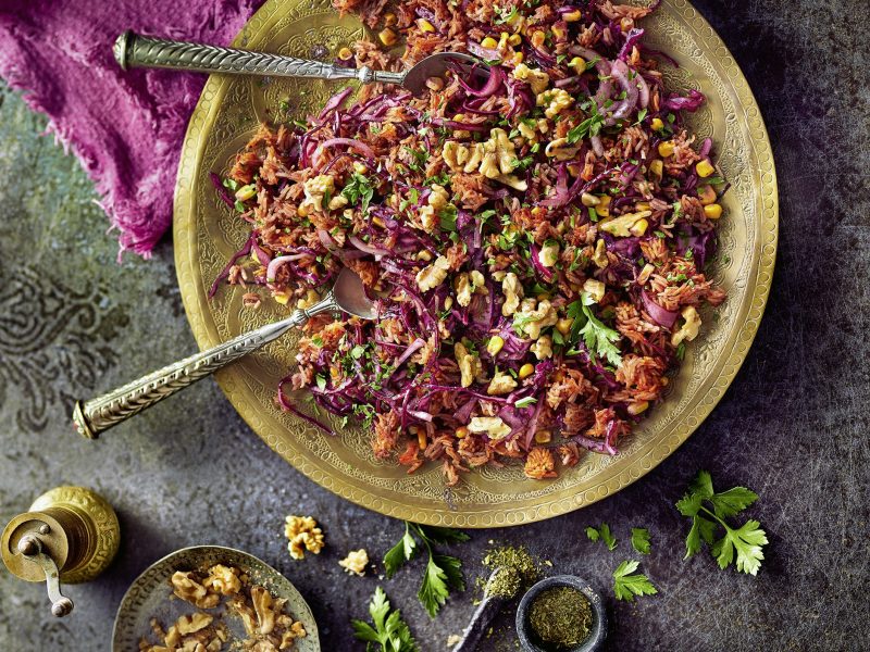 Ein hellgrüner Teller mit dem roten Reissalat auf einem grauen Tisch mit den dazugehörigen Gewürzen. Alles direkt von oben fotografiert.