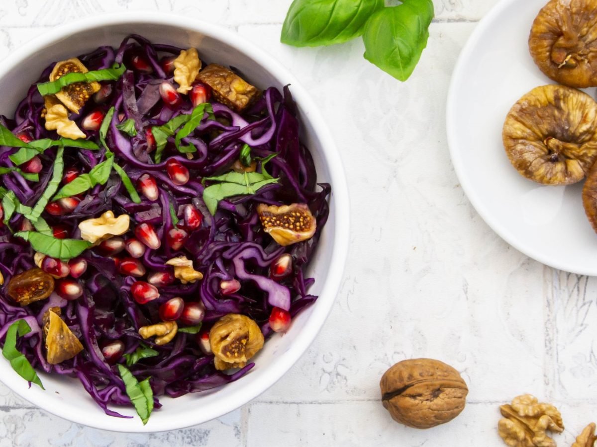 Rotkohl-Salat mit getrockneten Feigen in einer weißen Schüssel. Rechts davon ein kleiner Teller mit getrockneten Feigen. Auf dem weißen Hintergrund Basilikum verteilt.