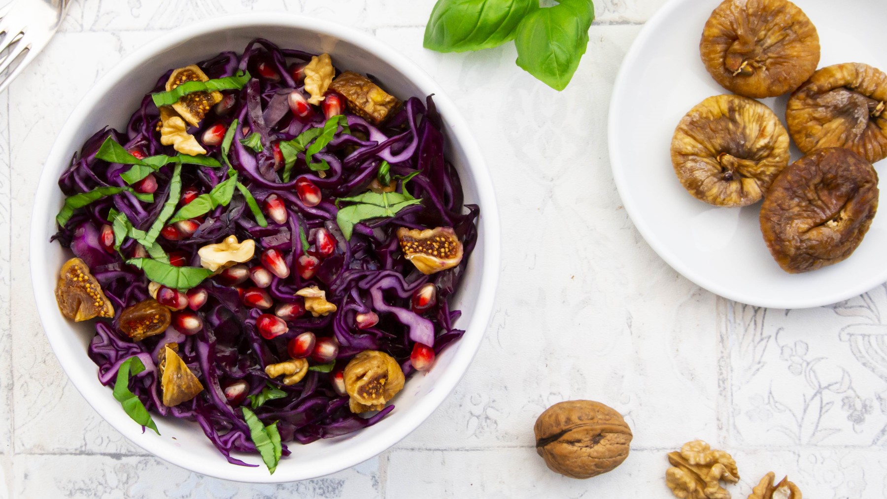 Rotkohl-Salat mit getrockneten Feigen in einer weißen Schüssel. Rechts davon ein kleiner Teller mit getrockneten Feigen. Auf dem weißen Hintergrund Basilikum verteilt.