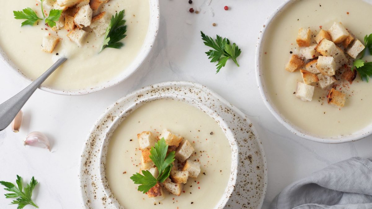 Drei Schalen cremige Knoblauchsuppe, garniert mit knusprigen Croutons und Petersilienblättern, auf einem weißen Untergrund mit Knoblauchzehen.