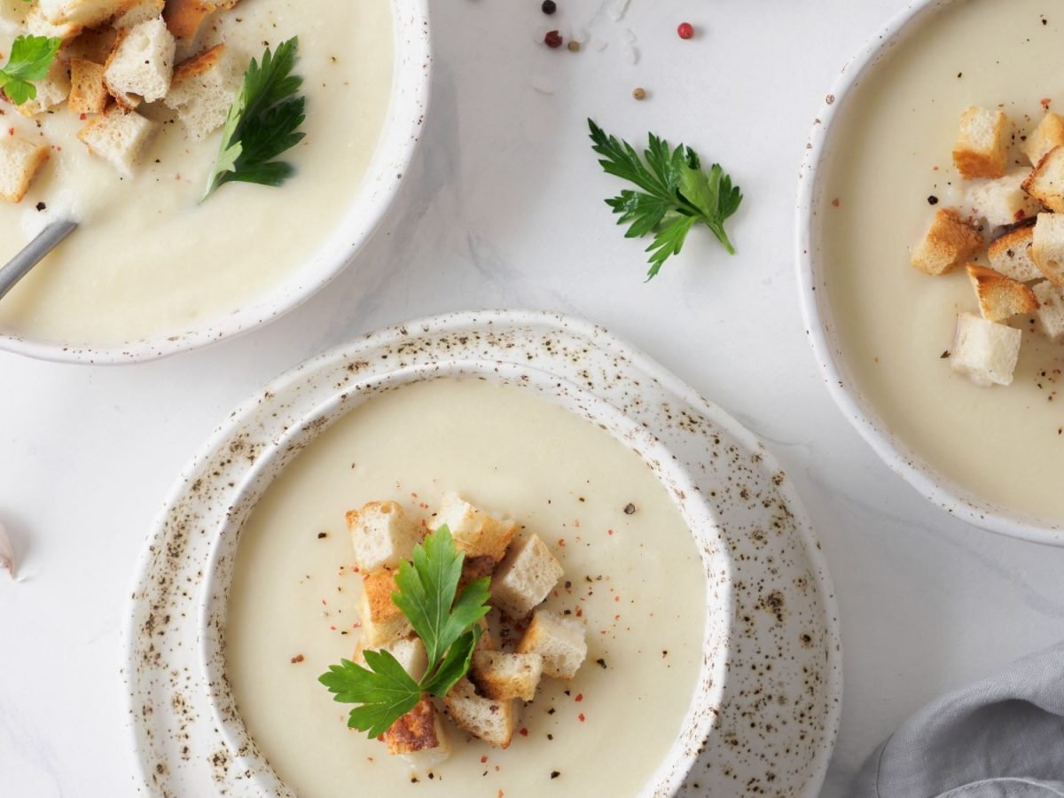 Drei Schalen cremige Knoblauchsuppe, garniert mit knusprigen Croutons und Petersilienblättern, auf einem weißen Untergrund mit Knoblauchzehen.