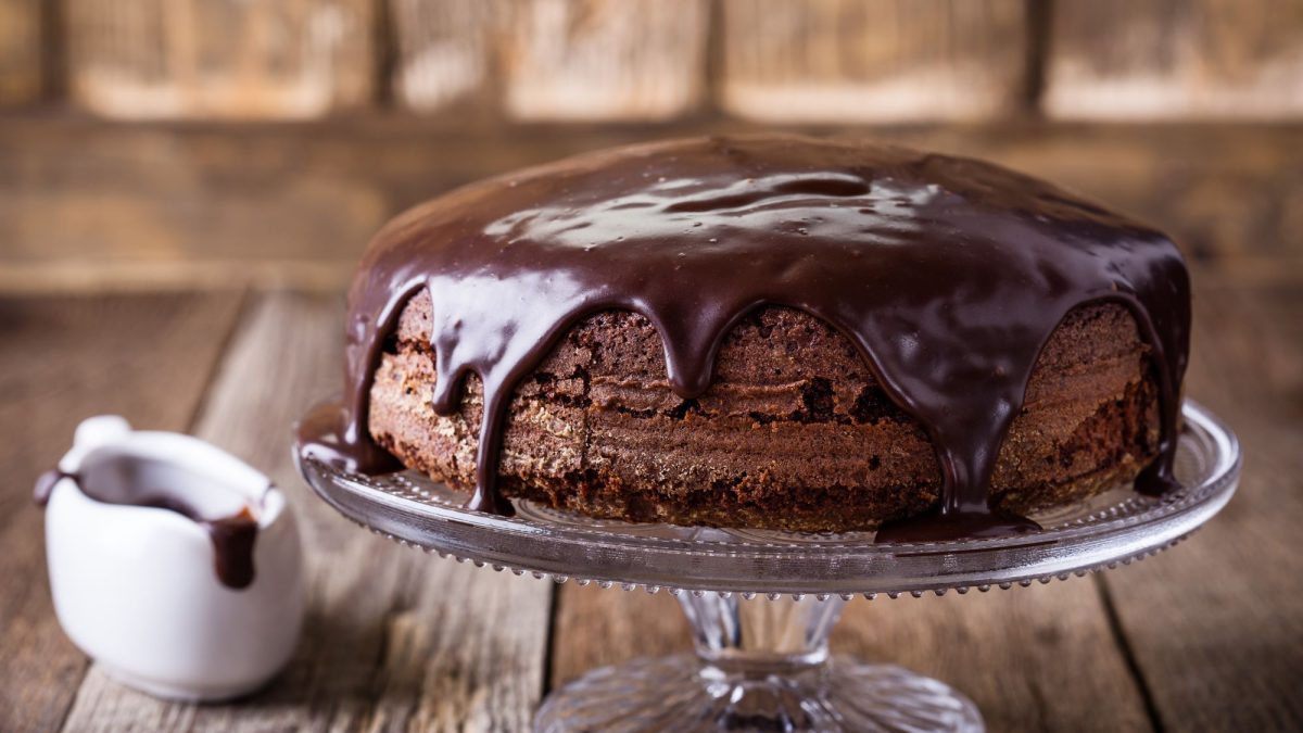 Saftiger Schokoladenkuchen mit glänzender Schokoglasur auf Glasplatte, daneben ein kleines Kännchen mit geschmolzener Schokolade auf rustikalem Holztisch.