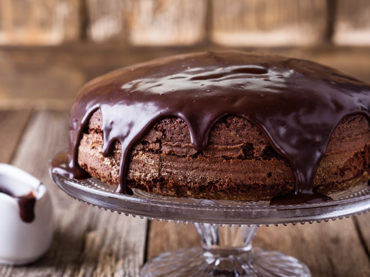 Saftiger Schokoladenkuchen mit glänzender Schokoglasur auf Glasplatte, daneben ein kleines Kännchen mit geschmolzener Schokolade auf rustikalem Holztisch.