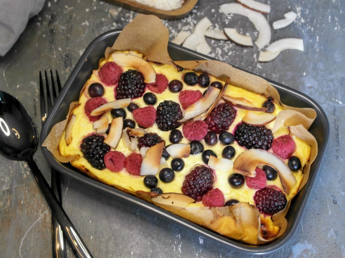 Ein ekleien graue auflaufform mit dem Skyr-Auflauf udn vielen beeren von schräg oben auf eienm grauen Steintisch fotografiert. Im Hintergrund ein graues Tuch und Kokoschips.