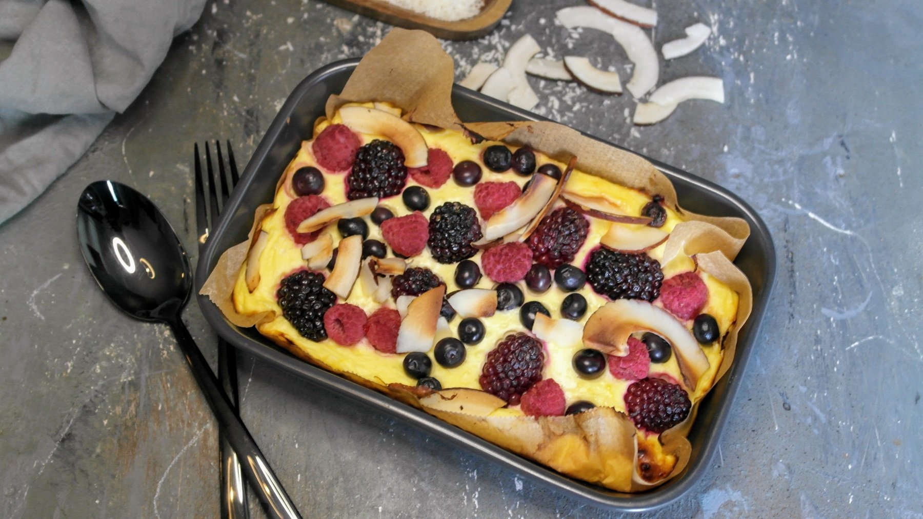 Ein ekleien graue auflaufform mit dem Skyr-Auflauf udn vielen beeren von schräg oben auf eienm grauen Steintisch fotografiert. Im Hintergrund ein graues Tuch und Kokoschips.