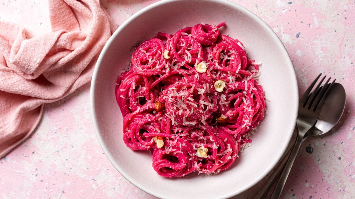 Pinke Spaghetti mit geriebenem Parmesan und Haselnüssen in einer weißen Schüssel auf pastellfarbenem Untergrund mit rosa Stoffserviette.