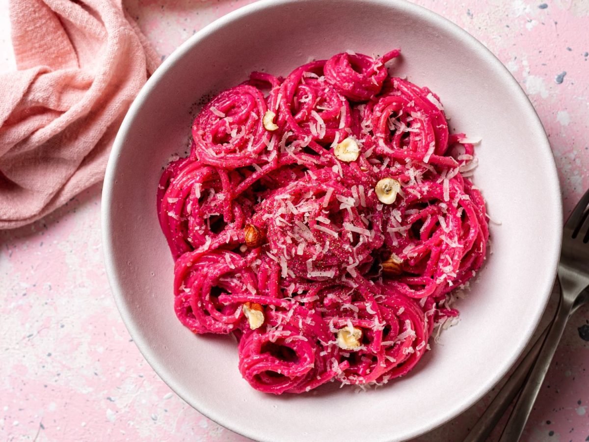 Pinke Spaghetti mit geriebenem Parmesan und Haselnüssen in einer weißen Schüssel auf pastellfarbenem Untergrund mit rosa Stoffserviette.