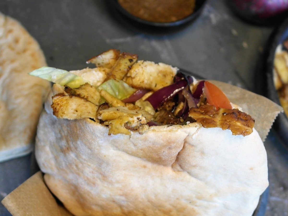 Ein gefülltes Pita-Brot, mit dem veganen Tofu-Gyros, Salat und Gurken gefüllt. Alles auf einem Steintisch, mit Gewürzen und Brot im Hintergrund.