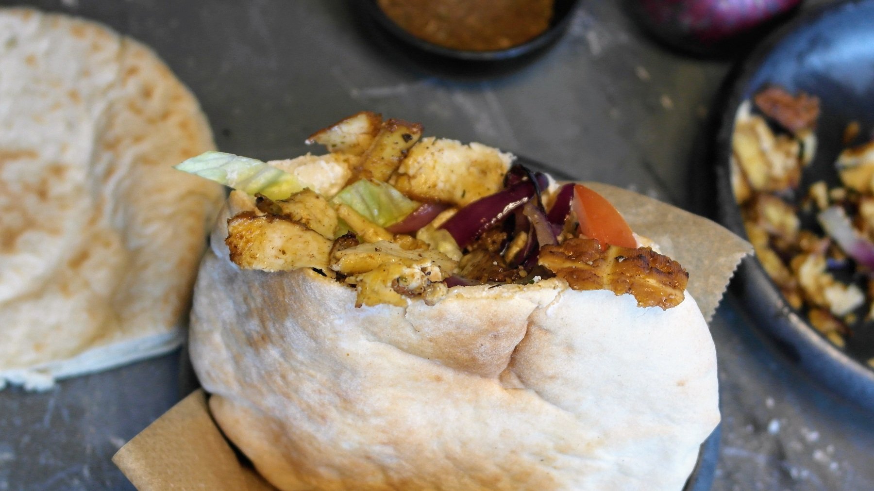 Ein gefülltes Pita-Brot, mit dem veganen Tofu-Gyros, Salat und Gurken gefüllt. Alles auf einem Steintisch, mit Gewürzen und Brot im Hintergrund.