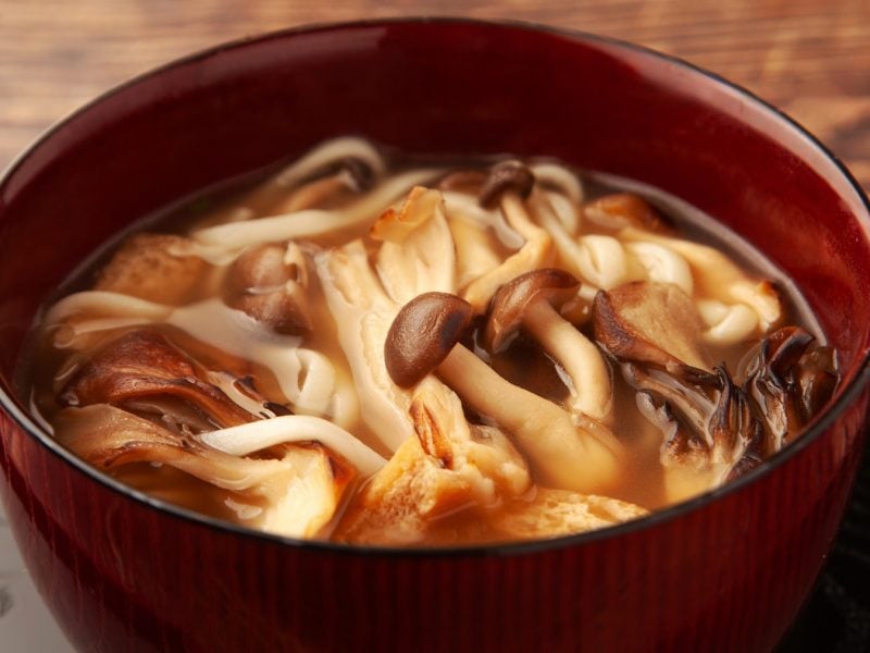 Eine braune Schale mit den Udon-Nudeln und Pilzen auf einem dunklen Holztablett und braunem Holztisch.