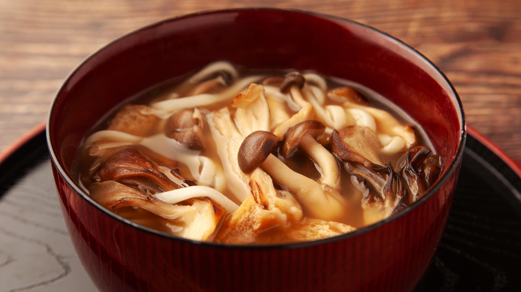 Eine braune Schale mit den Udon-Nudeln und Pilzen auf einem dunklen Holztablett und braunem Holztisch.