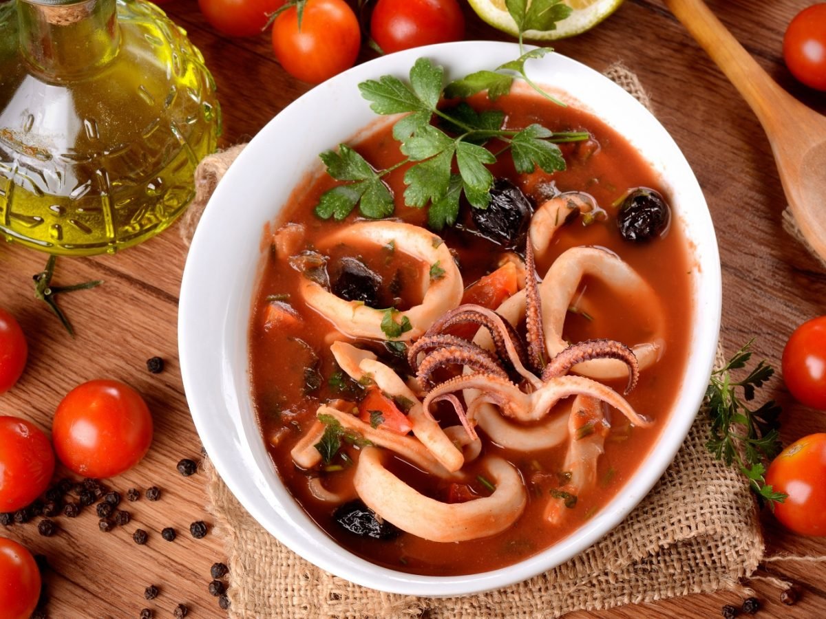 Draufsicht auf Zuppa di calamari, daneben frische Tomaten, Olivenöl und ein Holzkochlöffel.