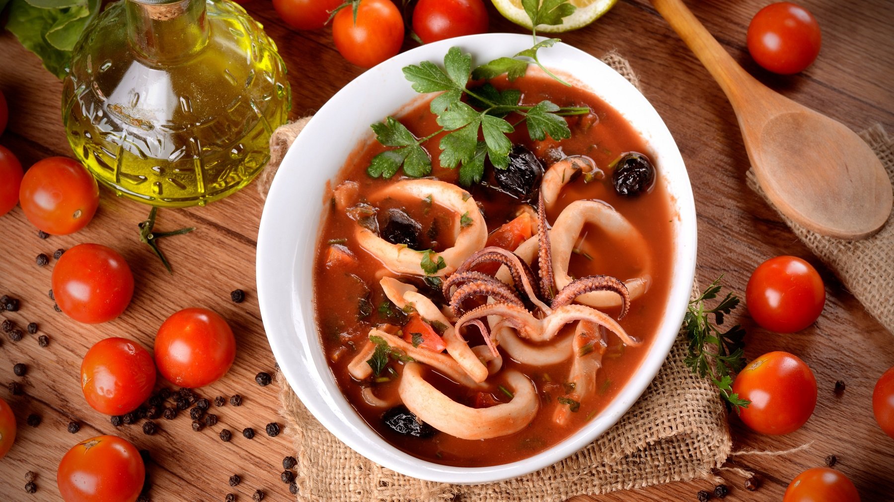 Draufsicht auf Zuppa di calamari, daneben frische Tomaten, Olivenöl und ein Holzkochlöffel.