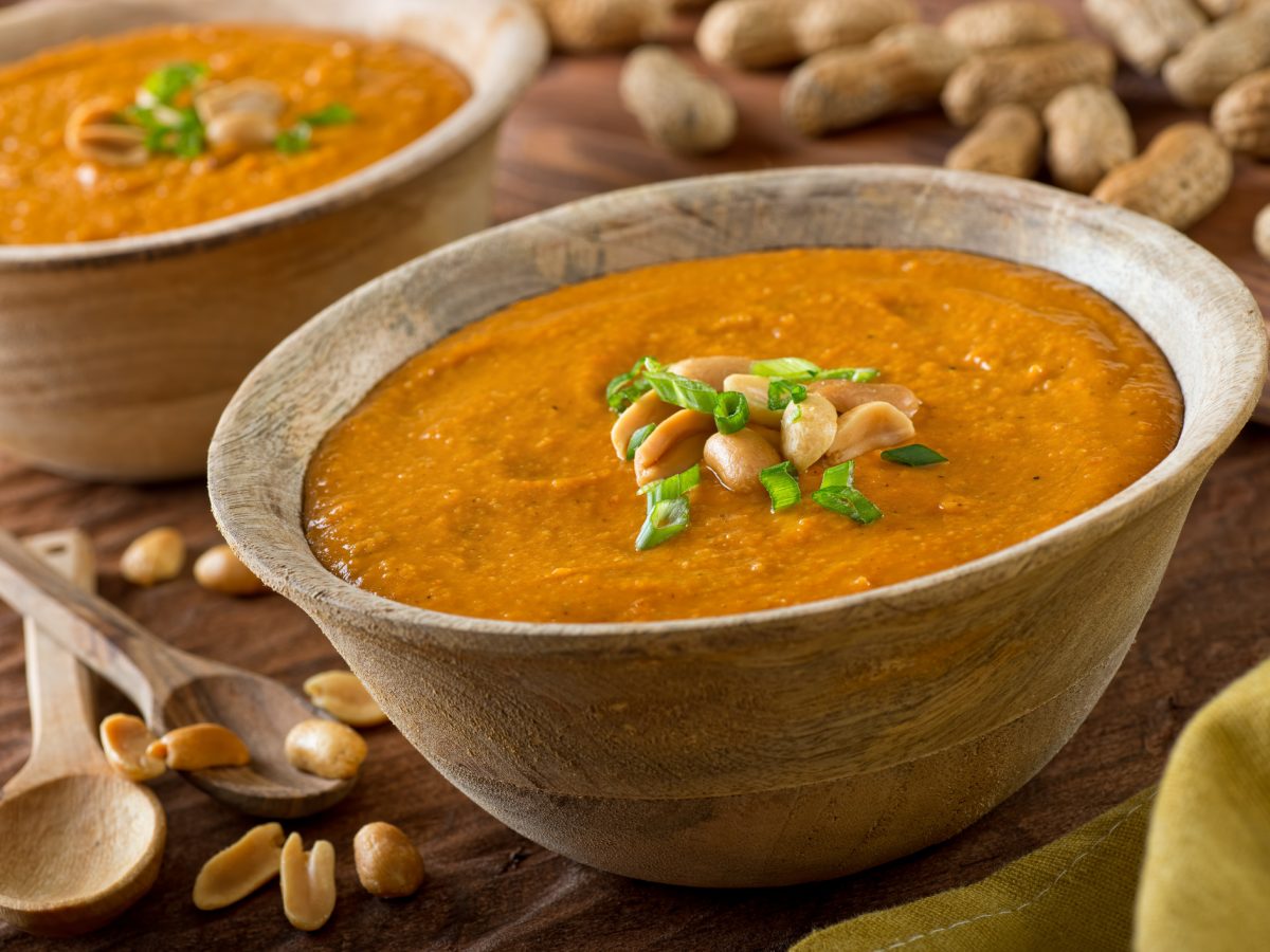 Zwei Schüsseln mit einer afrikanischen Erdnusssuppe auf einem rustikalen Untergrund aus Holz. Drum herum liegen frische Erdnüsse und zwei Löffel.