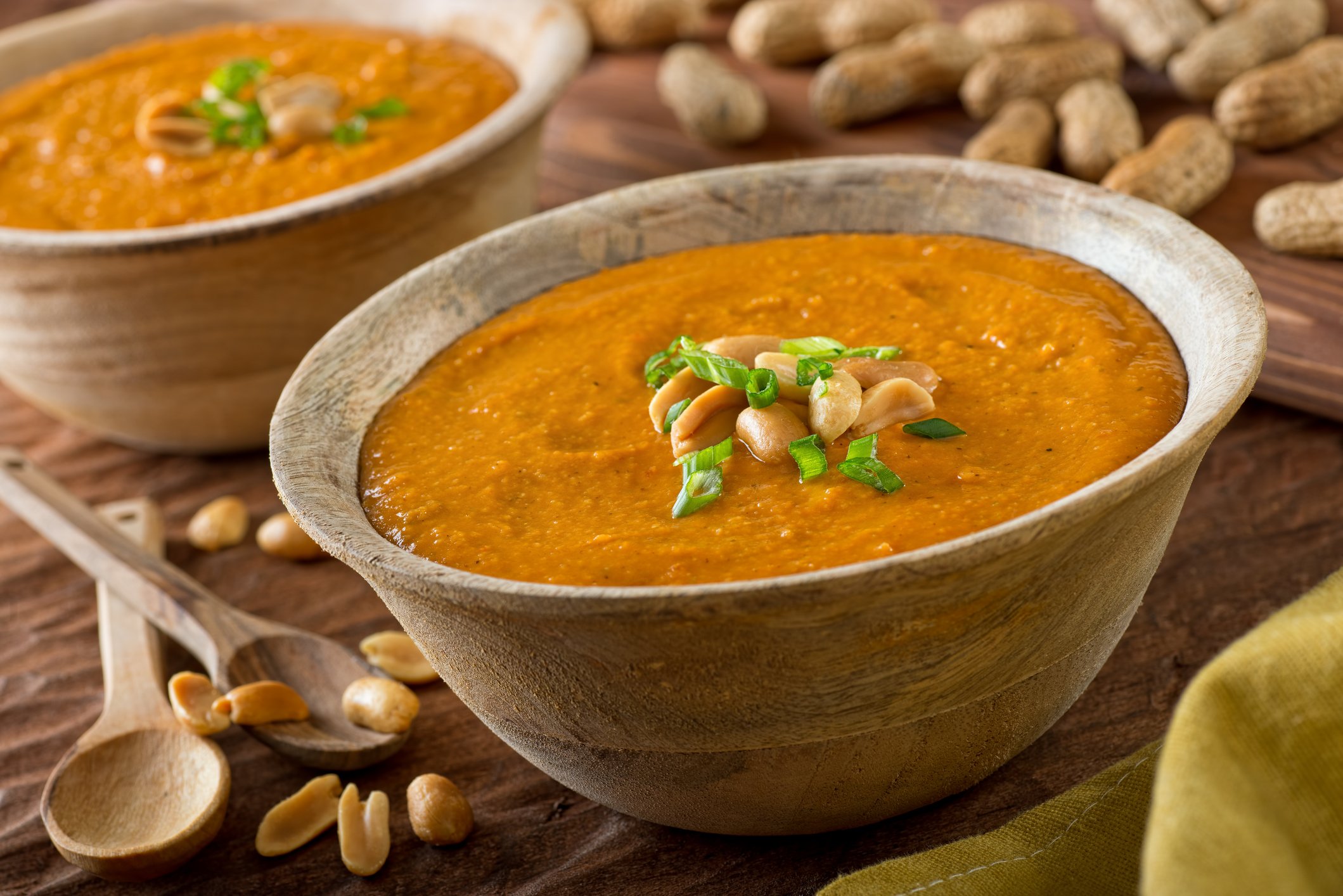 Zwei Schüsseln mit einer afrikanischen Erdnusssuppe auf einem rustikalen Untergrund aus Holz. Drum herum liegen frische Erdnüsse und zwei Löffel.