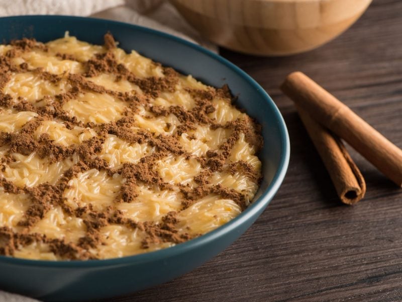 Eine Schale mit portugiesischem Aletria-Dessert, bestehend aus Fadennudeln in einer süßen, cremigen Masse, die mit Zimt in einem dekorativen Muster bestreut ist. Daneben liegen zwei Zimtstangen zur Dekoration.