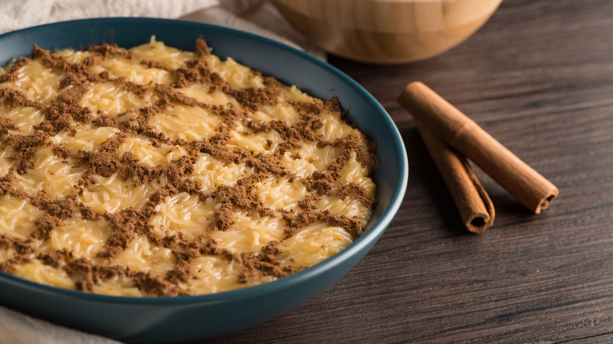 Eine Schale mit portugiesischem Aletria-Dessert, bestehend aus Fadennudeln in einer süßen, cremigen Masse, die mit Zimt in einem dekorativen Muster bestreut ist. Daneben liegen zwei Zimtstangen zur Dekoration.