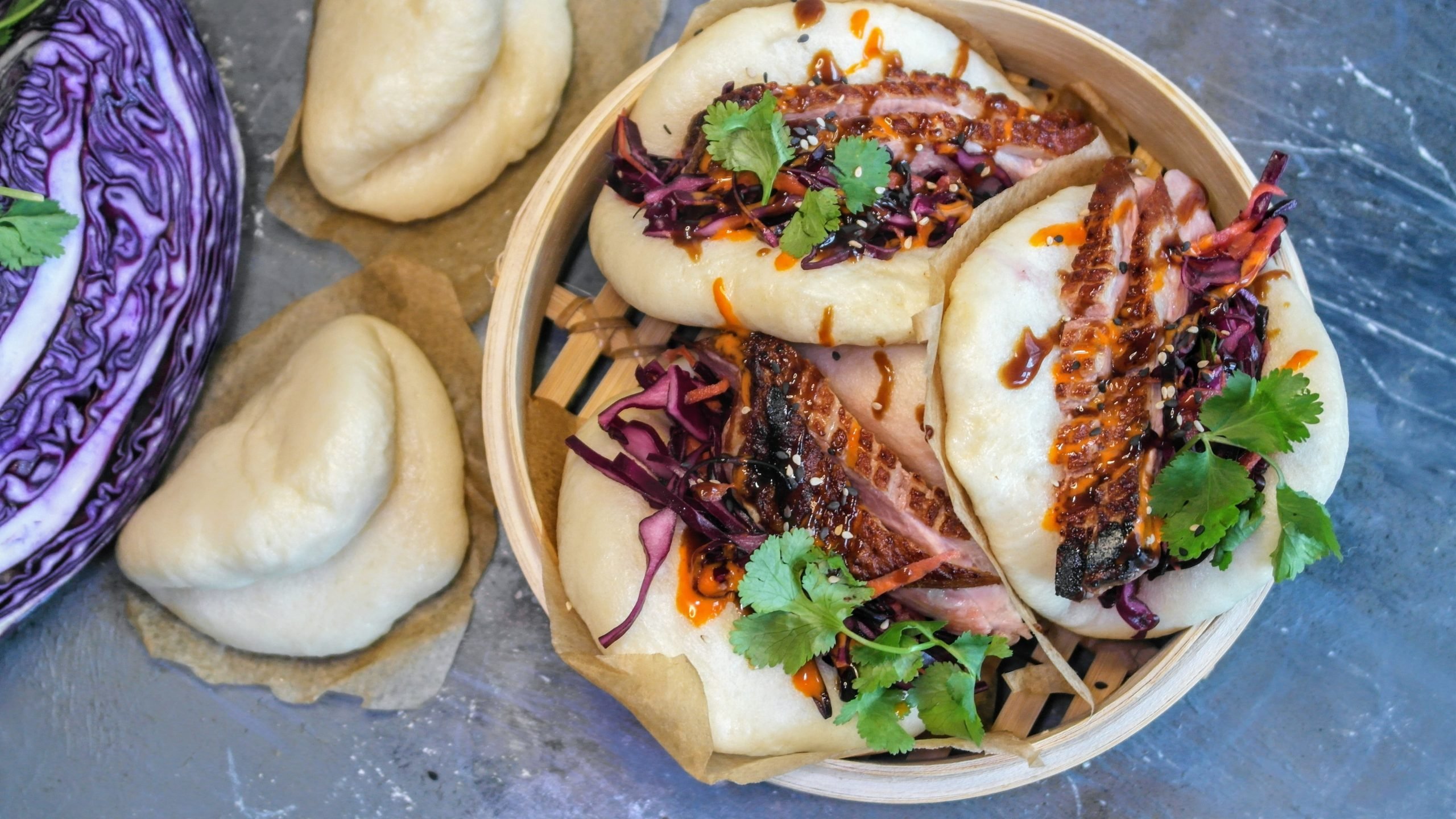 Ein Dampfkorb mit den Bao-Buns gefüllt auf einem grauen Steintisch. Daneben angeschnittener Rotkohl und ungefüllte Dampfbrötchen. Alles direkt von oben fotografiert.