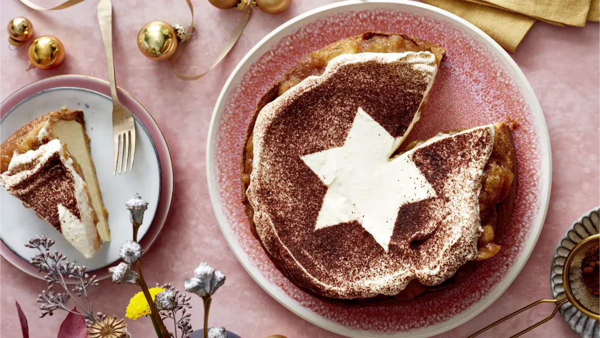 Bratapfel-Käsekuchen auf einem rosa Hintergrund. Links ein kleiner Teller mit einem Stück angerichtet und goldene Weihnachtskugeln verteilt.