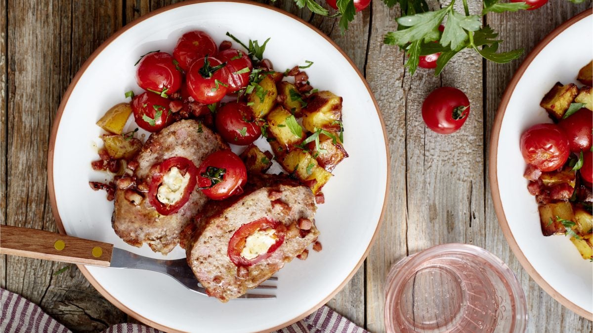 Draufsicht: Holzfäller-Braten mit Paprika in der Mitte auf einem Teller mit Ofentomaten- und Kartoffeln. Holzuntergrund.