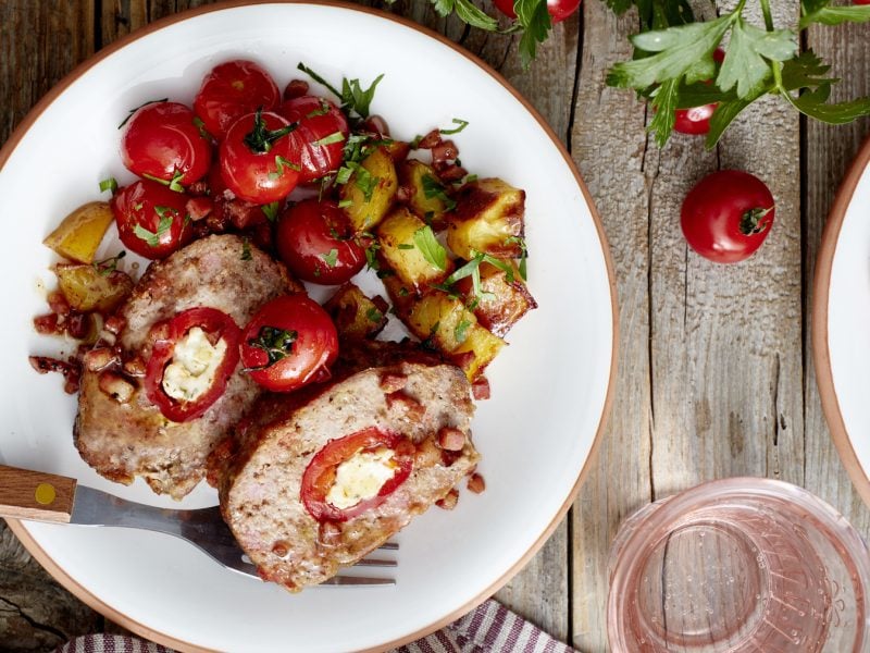 Draufsicht: Holzfäller-Braten mit Paprika in der Mitte auf einem Teller mit Ofentomaten- und Kartoffeln. Holzuntergrund.