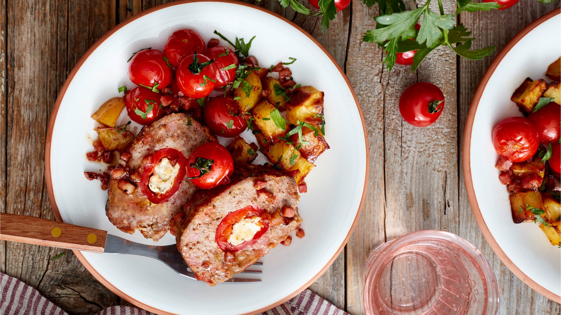 Draufsicht: Holzfäller-Braten mit Paprika in der Mitte auf einem Teller mit Ofentomaten- und Kartoffeln. Holzuntergrund.