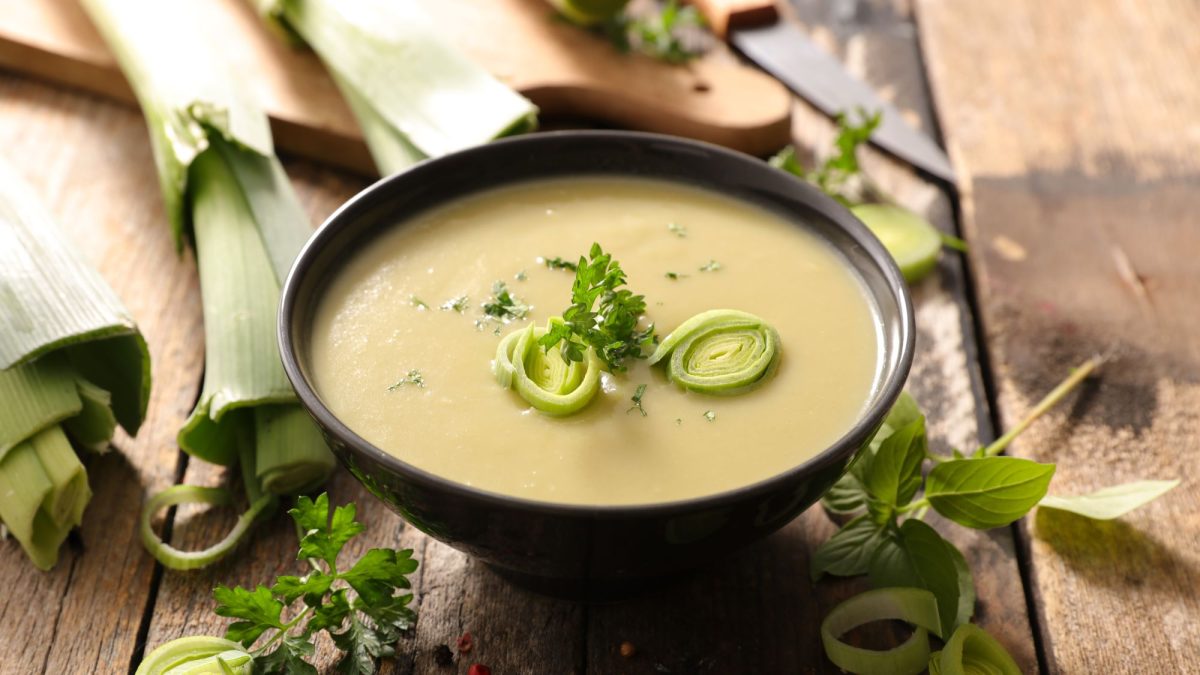 Eine Schale mit cremiger Lauchsuppe (Cawl Cennin), die mit frischen Kräutern und fein geschnittenen Lauchringen dekoriert ist. Im Hintergrund liegt frischer Lauch auf einem rustikalen Holzuntergrund.