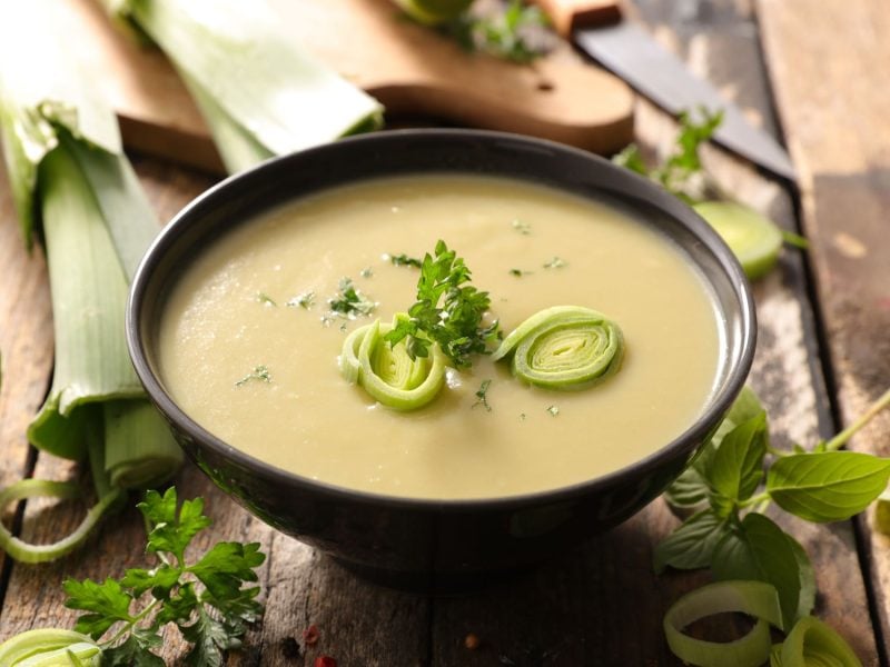 Eine Schale mit cremiger Lauchsuppe (Cawl Cennin), die mit frischen Kräutern und fein geschnittenen Lauchringen dekoriert ist. Im Hintergrund liegt frischer Lauch auf einem rustikalen Holzuntergrund.