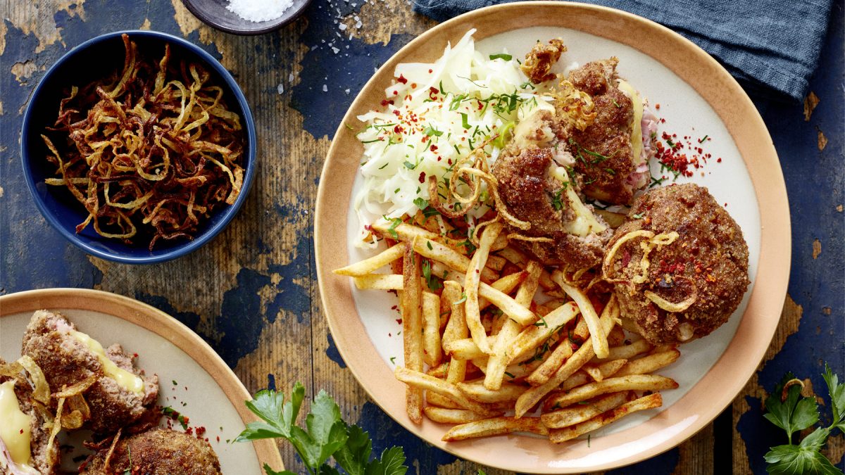 Draúfsicht: Cordon-bleu-Frikadellen mit Pommes auf einem Teller, daneben Schale mit Röstzwiebeln auf blauem Holzuntergrund.