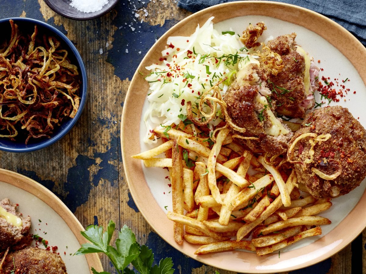 Draúfsicht: Cordon-bleu-Frikadellen mit Pommes auf einem Teller, daneben Schale mit Röstzwiebeln auf blauem Holzuntergrund.