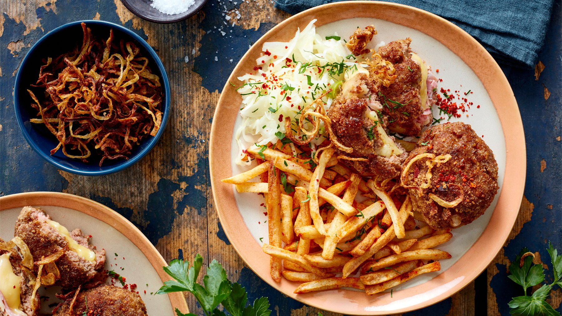 Draúfsicht: Cordon-bleu-Frikadellen mit Pommes auf einem Teller, daneben Schale mit Röstzwiebeln auf blauem Holzuntergrund.