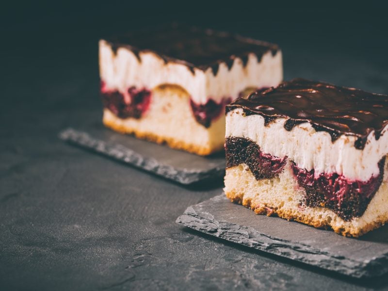 Zwei saftige Donauwellen-Stücke mit heller Creme, Kirschfüllung und glänzendem Schokoladenguss auf dunklen Schieferplatten.