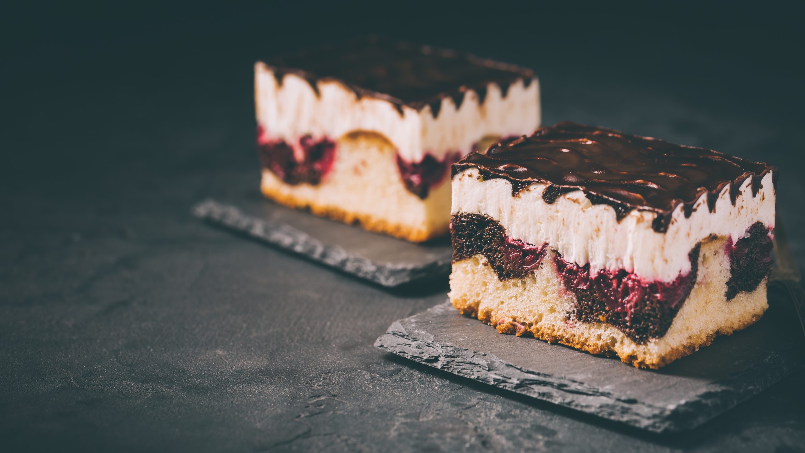 Zwei saftige Donauwellen-Stücke mit heller Creme, Kirschfüllung und glänzendem Schokoladenguss auf dunklen Schieferplatten.