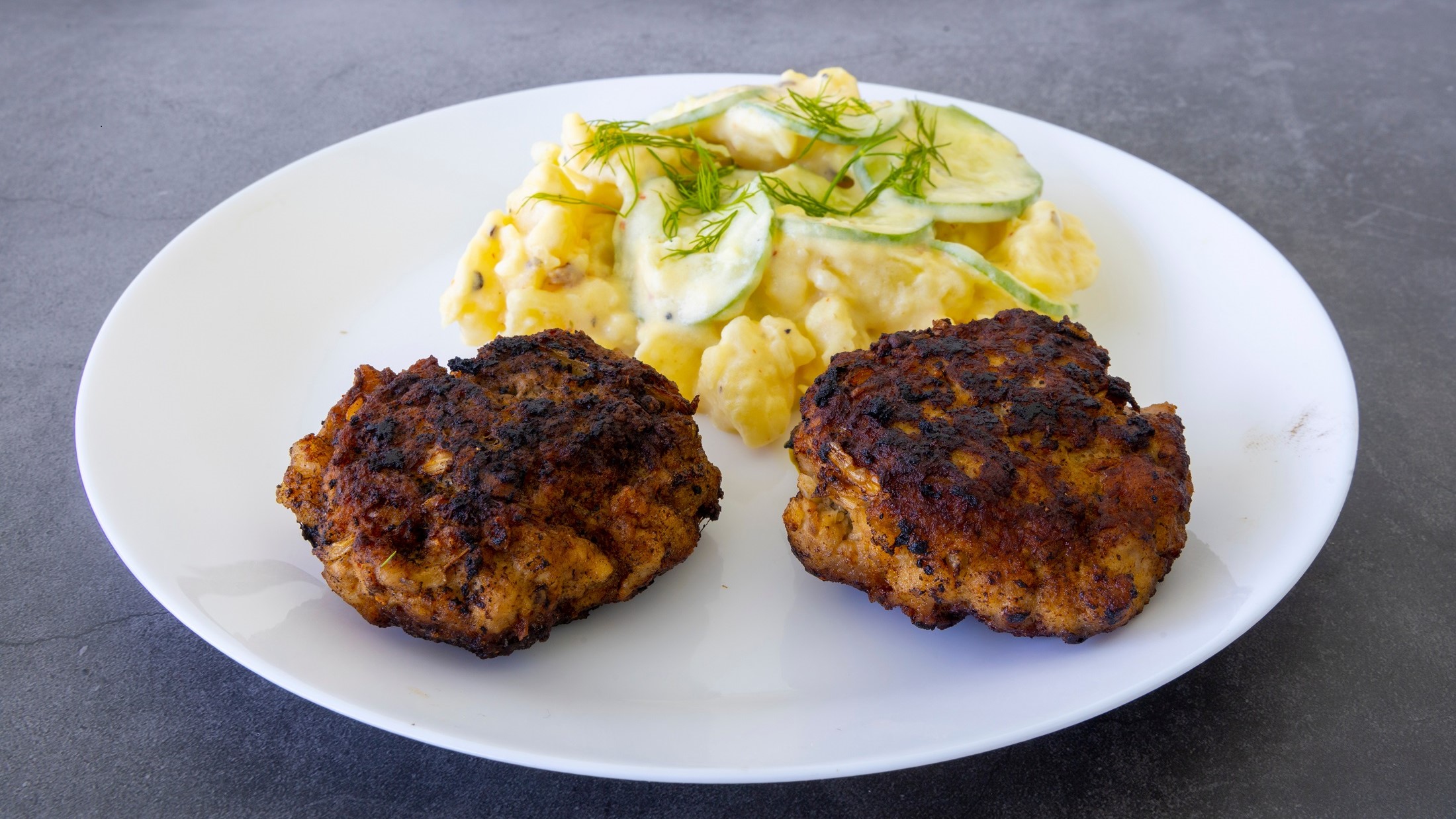 Berliner Buletten mit Kartoffelsalat und Gurken auf einem weißen Teller. Draufsicht schräg