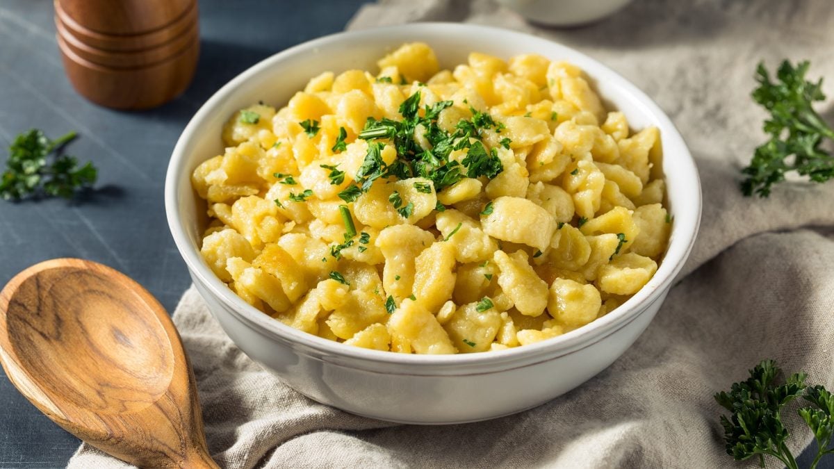 Draufsicht, schräg: hausgemachte Eierspätzle in einer weißen Schüssel. Sie stehen auf einem Tisch. Drum herum sind ein Holzlöffel, sowie der Fuß einer Pfeffermühle zu sehen.