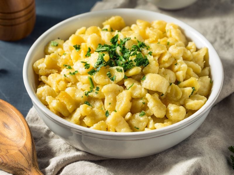 Draufsicht, schräg: hausgemachte Eierspätzle in einer weißen Schüssel. Sie stehen auf einem Tisch. Drum herum sind ein Holzlöffel, sowie der Fuß einer Pfeffermühle zu sehen.