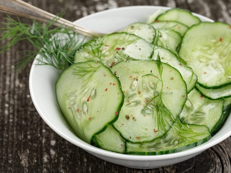 Gurkensalat mit Essig & Öl, Dill und rotem Pfeffer in einer weißen Salatschale. Die steht auf einem rustikalem Holzuntergrund.