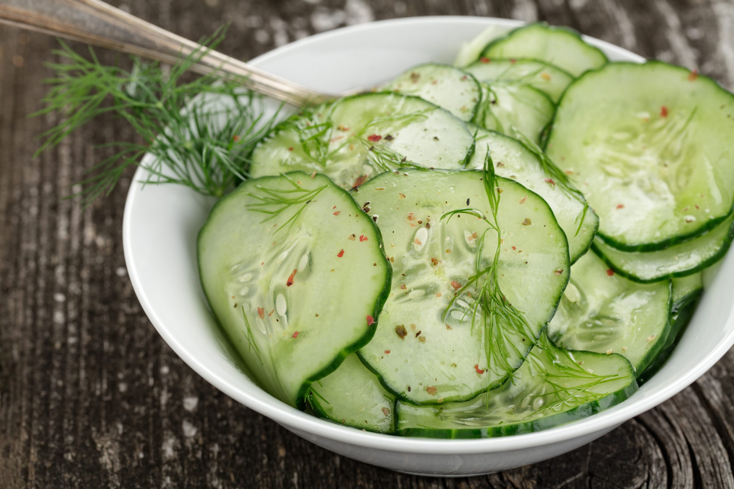 Gurkensalat mit Essig & Öl, Dill und rotem Pfeffer in einer weißen Salatschale. Die steht auf einem rustikalem Holzuntergrund.